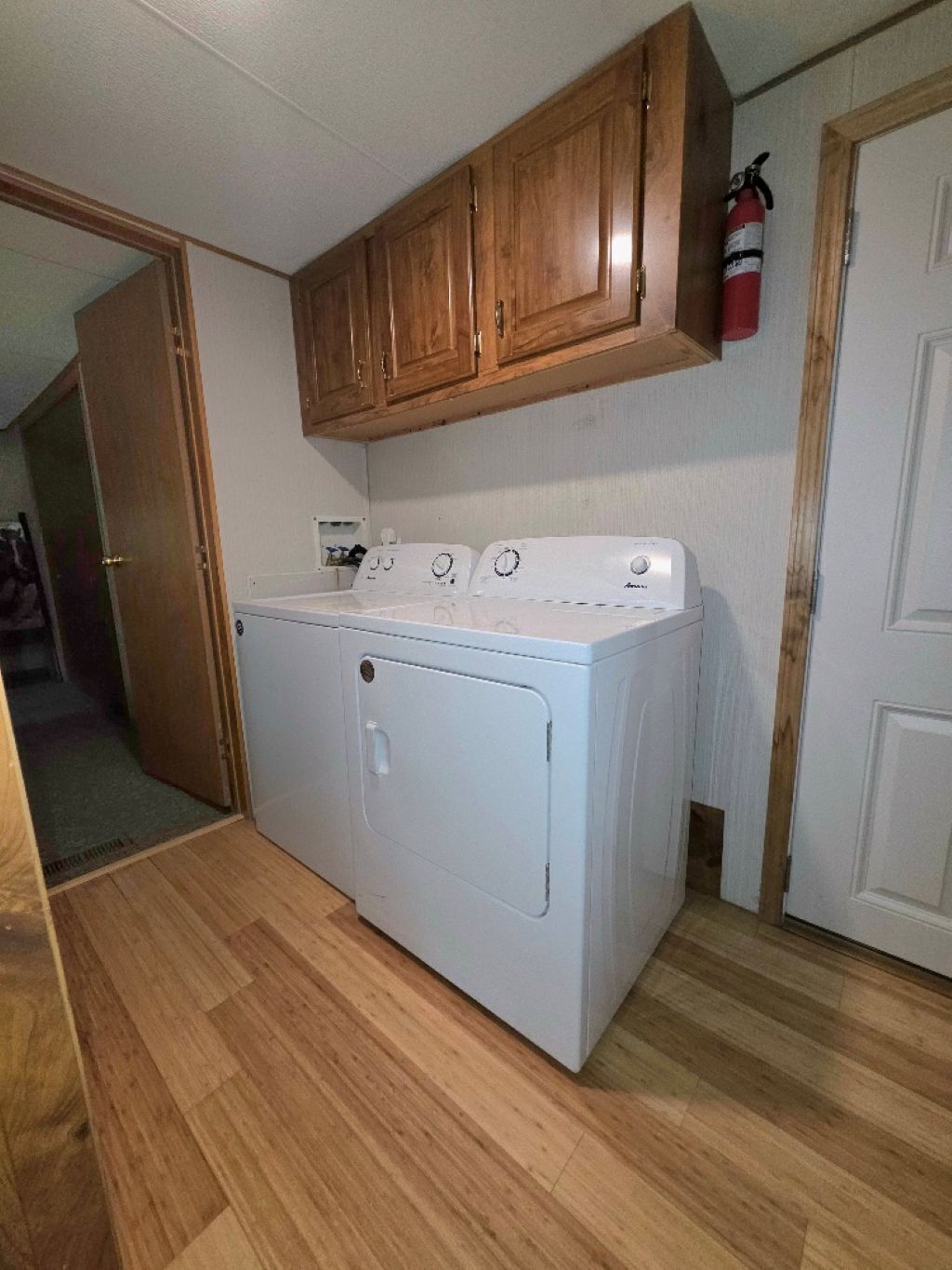 A compact laundry room with a front-loading washer and dryer, wooden cabinets above, and a fire extinguisher on the wall.