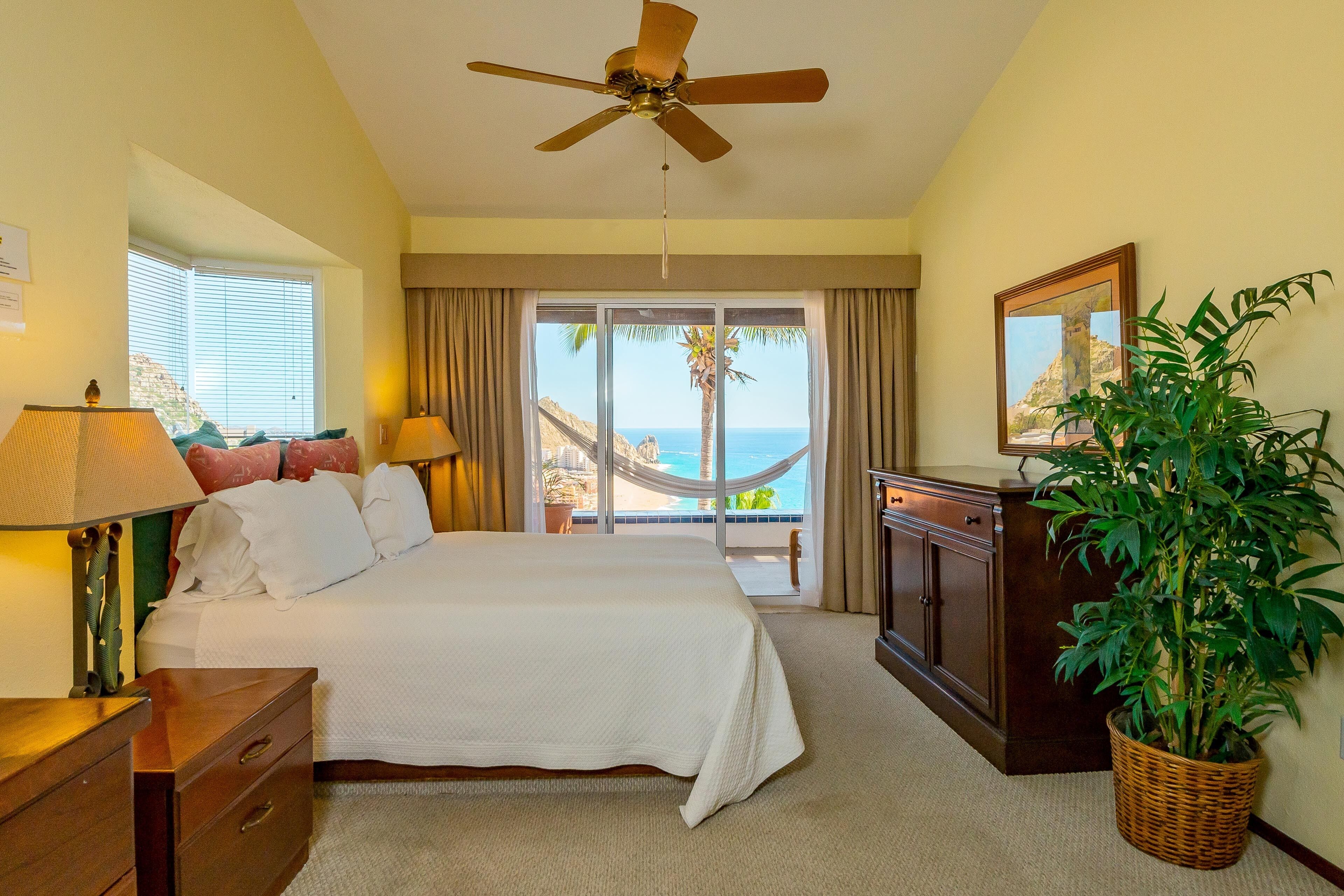 A hotel room with a bed, a ceiling fan, a nightstand, a lamp, a dresser, a potted plant, and a view of the ocean through a sliding door.