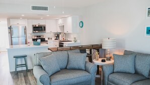 Eine moderne Küche mit weißen Schränken, Edelstahlgeräten und einer zentralen Kochinsel. Ein graues Sofa und ein kleiner runder Tisch mit einer Lampe sind ebenfalls vorhanden.