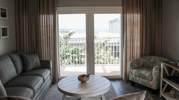 Ein Zimmer mit einer Couch, einem Sessel, einem kleinen Tisch und Blick durch eine Schiebetür auf einen Balkon mit Pflanzen und ein Gebäude.