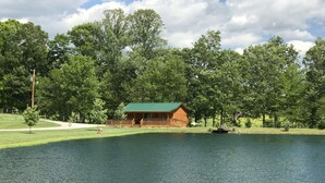 A lodge with a green roof, situated by a lake, surrounded by dense trees. - Private Catch & Release Lake - Boat Rental Included! Close To Lake Cumberland! (Russell Springs)