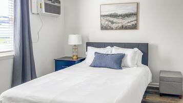 A hotel room with a bed, a nightstand with a lamp, a gray ottoman, a painting on the wall, and an air conditioning unit.