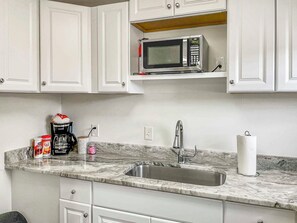 Una cocina con gabinetes blancos, un lavamanos de acero inoxidable, un microondas, una cafetera y un dispensador de toallas de papel.