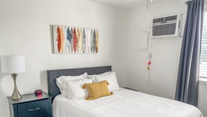 A neatly made bed with white bedding, a bedside lamp, a wall-mounted air conditioner, and a colorful abstract painting above the bed. - 2 BR 8 min to Downtown - Free Parking -C (Nashville)