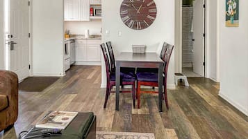 A dining area with a wooden table and chairs, a large wall clock, and a kitchen in the background.