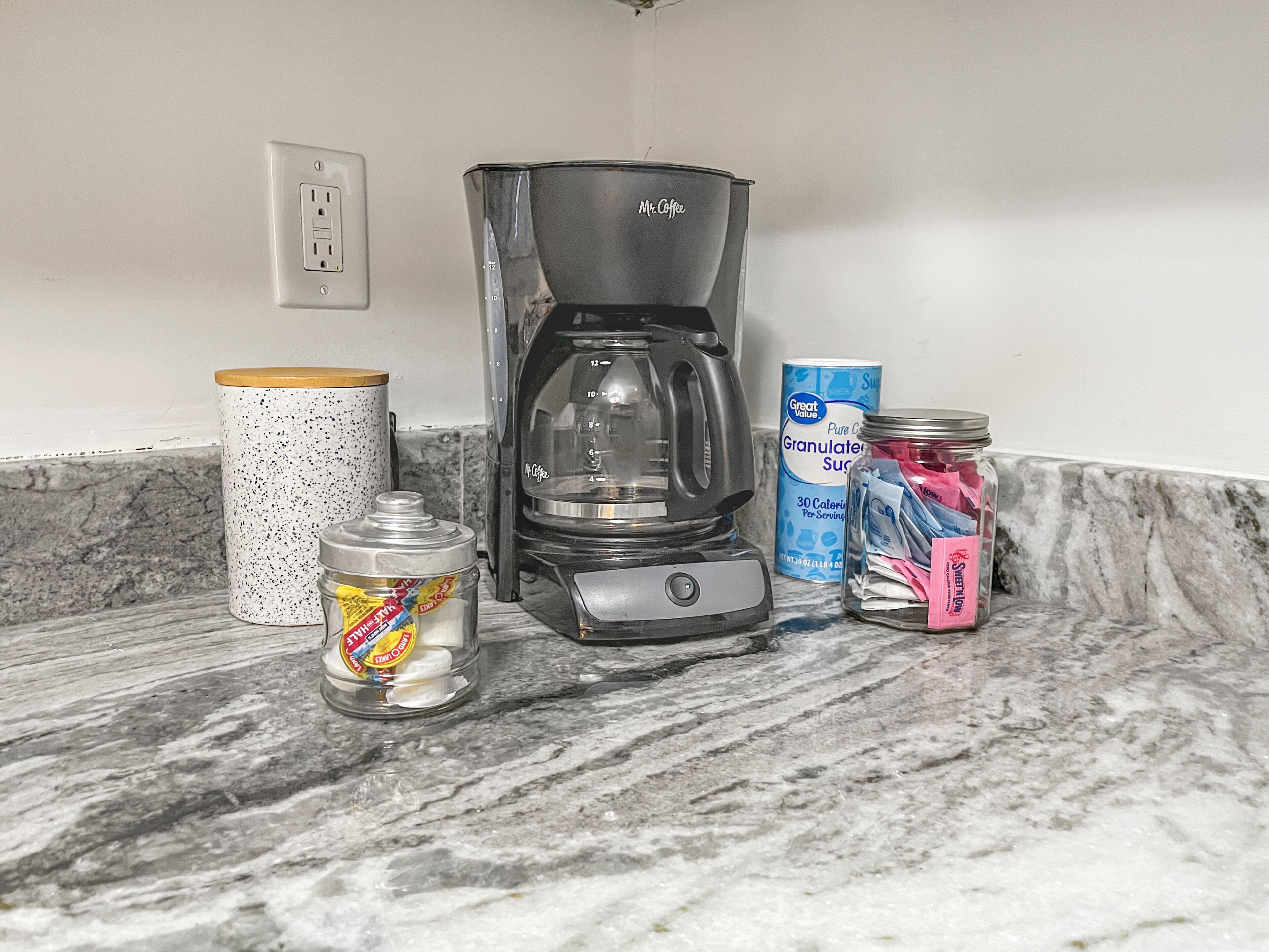 A kitchen counter with a coffee maker, a jar of sugar, a container of granulated sugar, and a box of tea bags.