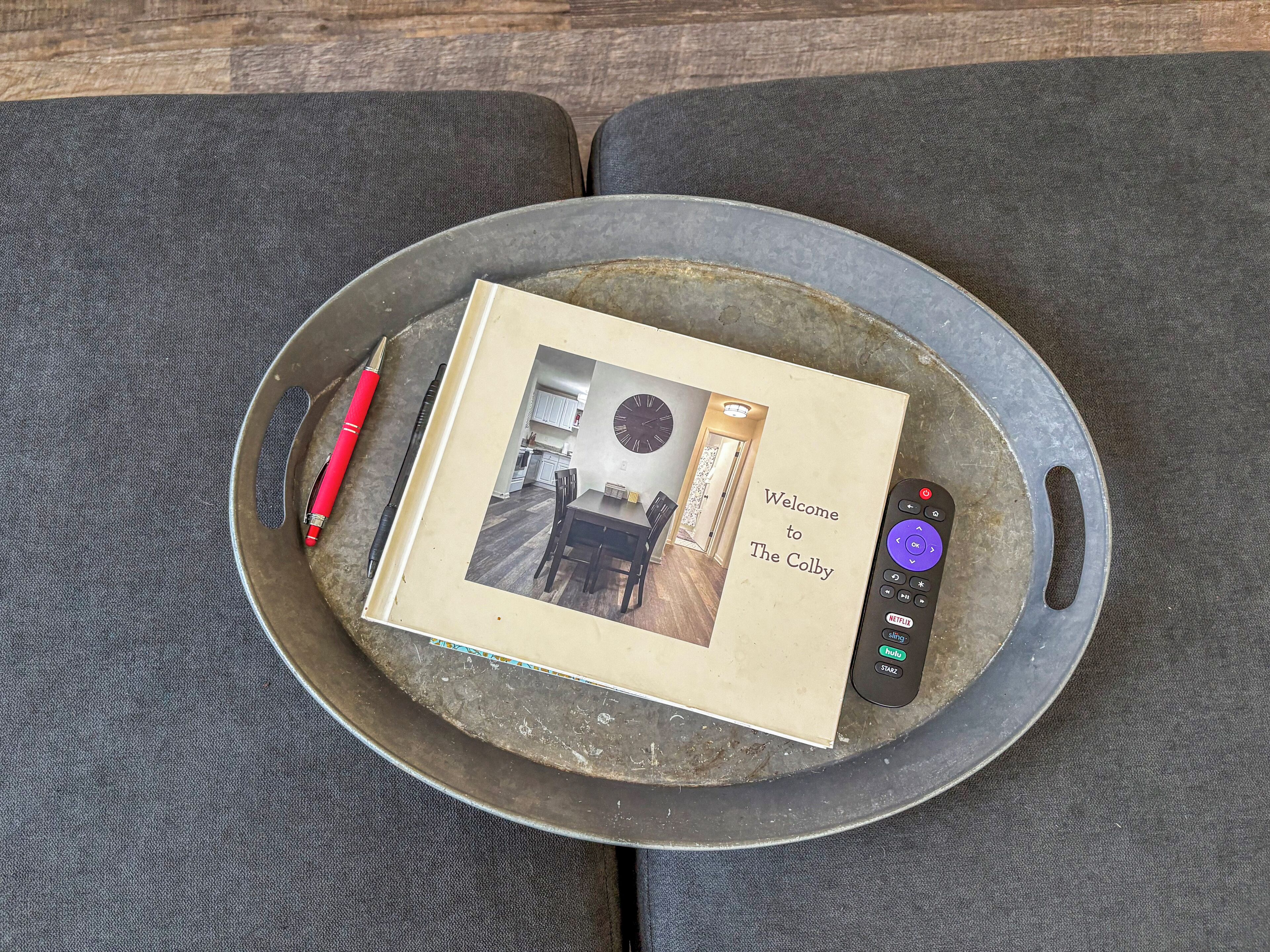 Un plateau contenant une télécommande, un stylo rouge et un livret intitulé « Bienvenue au Colby », posé sur un canapé gris.