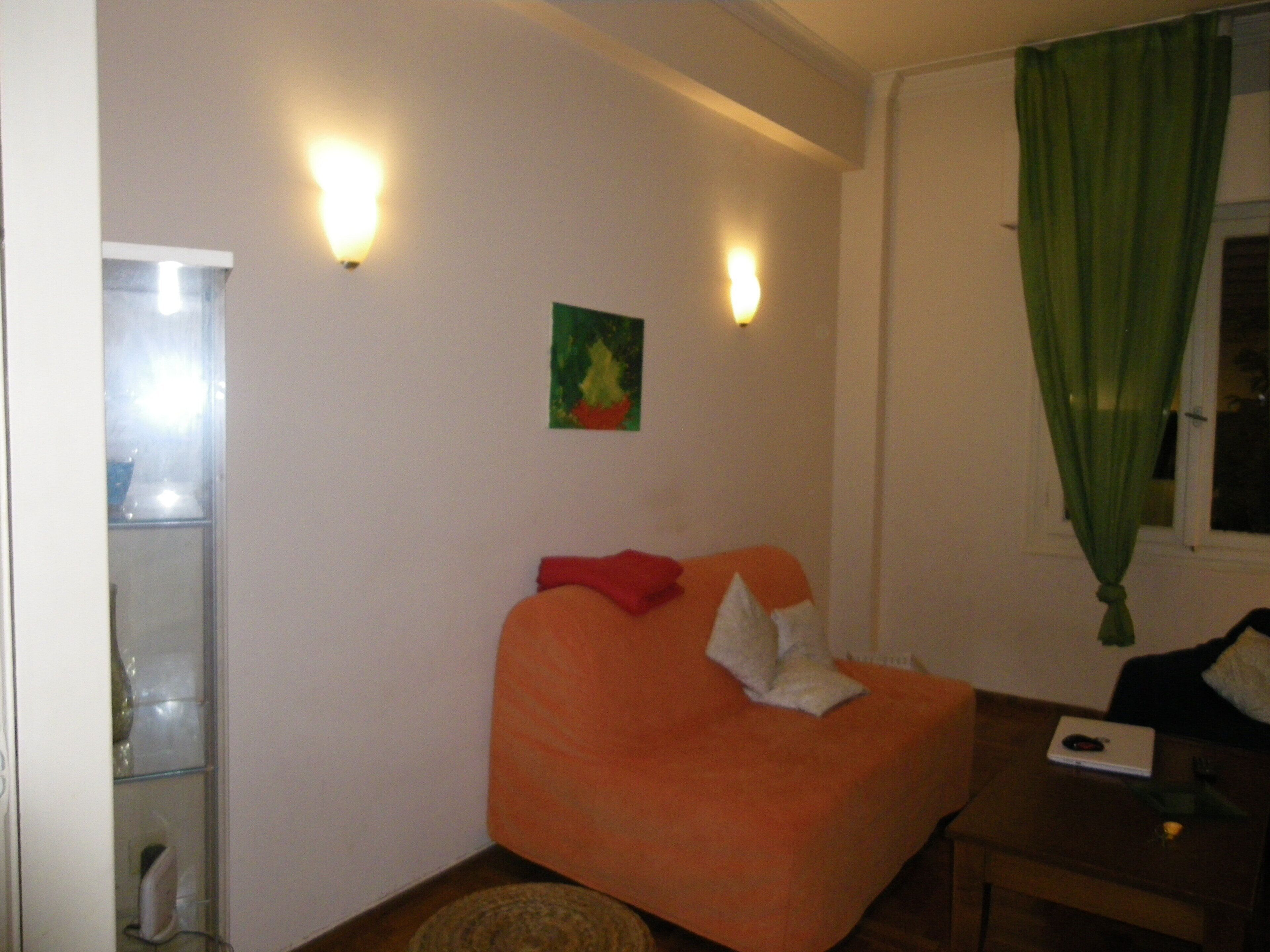 A small, orange, L-shaped sofa with a cushion and a pillow, a small table with a laptop, and a green curtain.