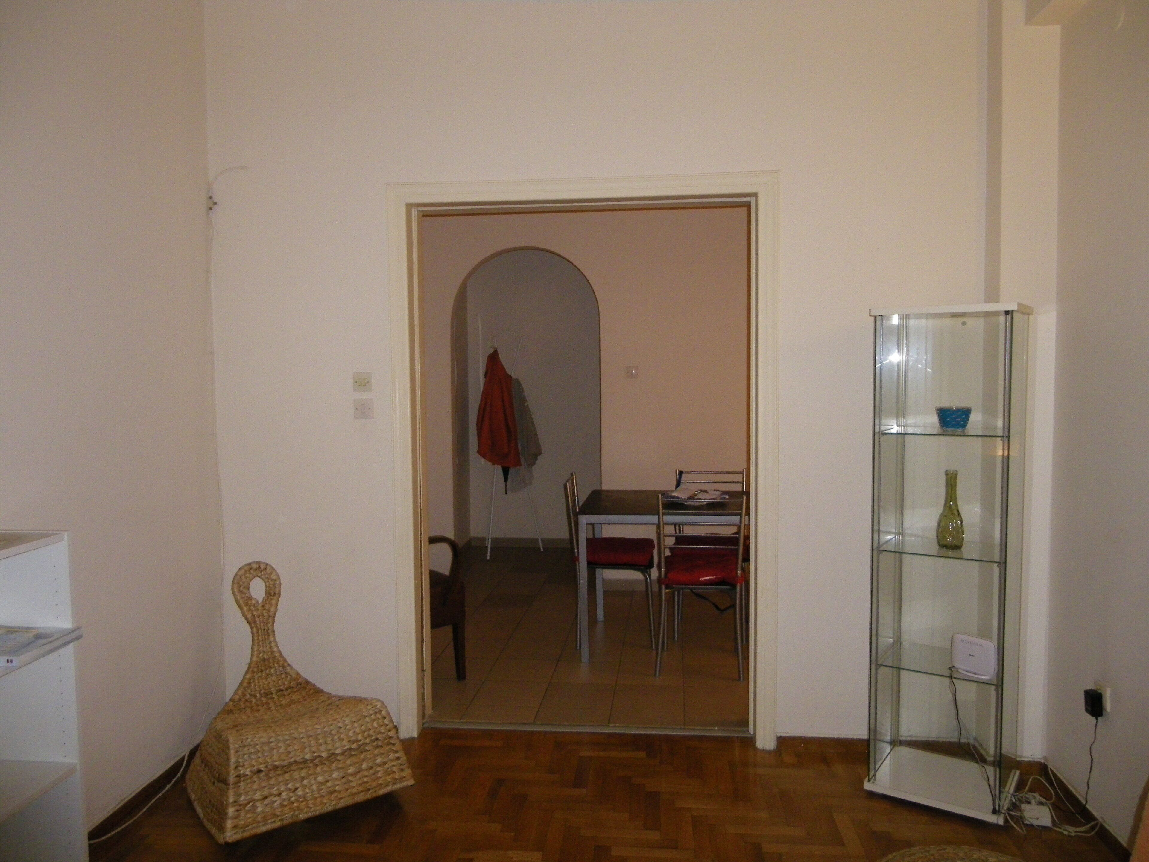 Une pièce avec un plancher en bois, une chaise en osier, une vitrine contenant un vase, et un coin repas avec des chaises et une table.