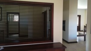 A room with a large window with blinds, a wooden desk, a chair, and a television. - The Moon and Sixpence Garden Guest House (Muldersdrift)