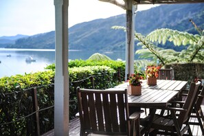 A wooden table with chairs and potted plants, overlooking a lake with boats and mountains in the background.