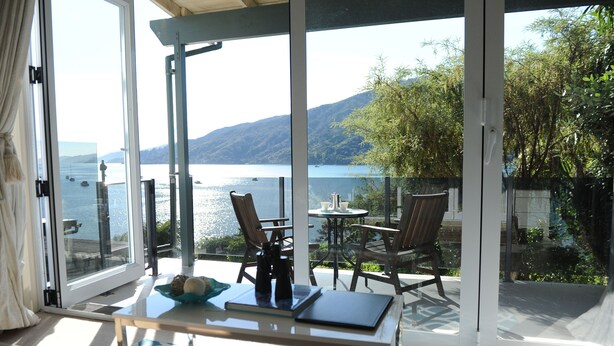 A room with a glass door leading to a balcony with a view of a lake and mountains. There is a table with chairs and a small table with a cup and saucer. - Tirimoana House (Anakiwa)