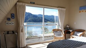 A bedroom with a large window, a bed, two wicker chairs, a small table, and a sign that reads "EXIT". - Tirimoana House (Anakiwa)