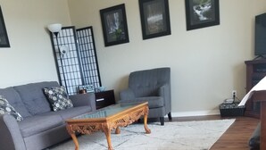 A living room with a grey sofa, a glass-top coffee table with carved legs, a grey armchair, a white rug, and framed pictures on the wall.