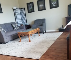 Una sala de estar con un sofá gris, una mesa de centro de cristal con patas talladas, un sillón gris, una alfombra blanca y cuadros enmarcados en la pared.