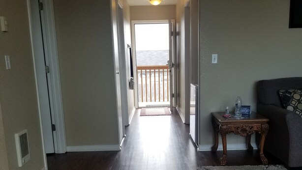 A hallway with a wooden floor leading to a balcony with a railing. There is a door on the left and a small table with a bottle and glass on the right.