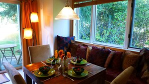 A dining area with a table set for two, a sofa with cushions, and a view of greenery through large windows. - Jackson Hole Condo, 5 Miles from Grand Teton National Park,4 miles to ski resort (Wilson)