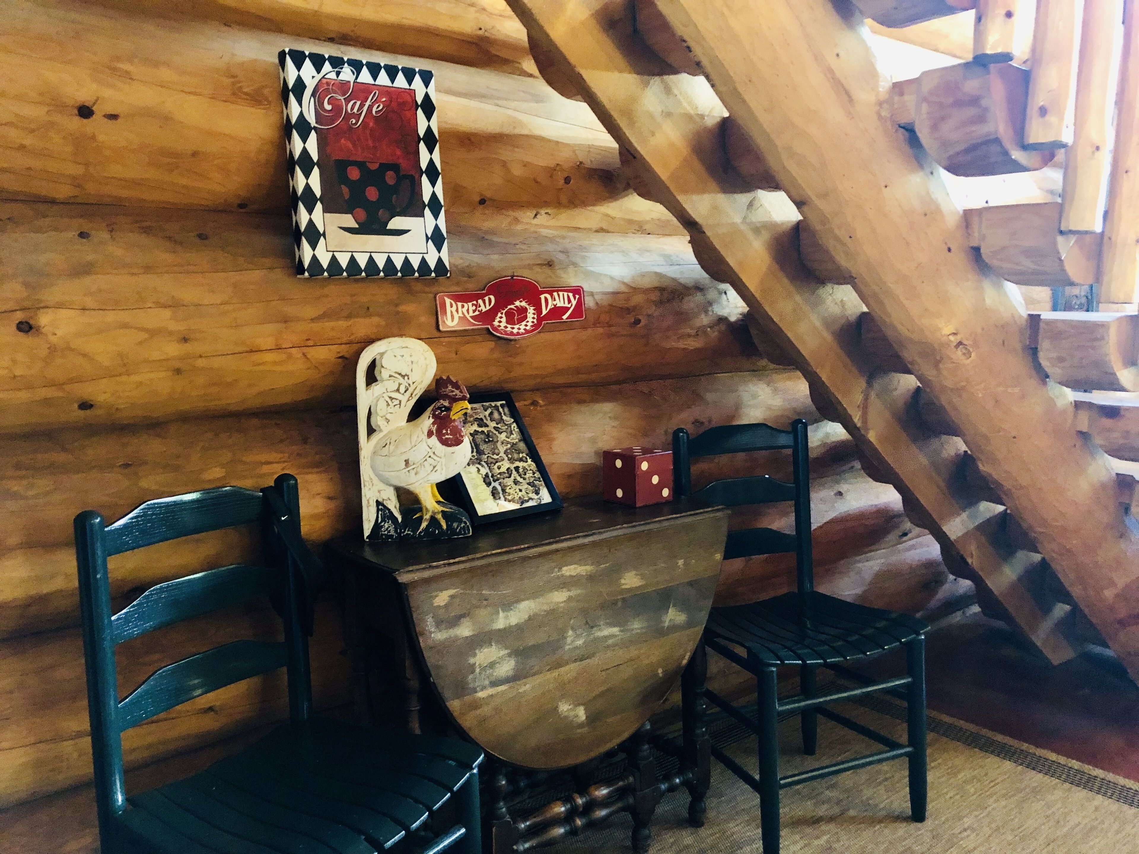 Un intérieur rustique en bois, avec une table en bois et deux chaises, un coq décoratif et une enseigne sur laquelle on peut lire « Pain frais tous les jours ».