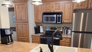 A kitchen with wooden cabinets, a stainless steel refrigerator, a microwave, an oven, a sink with a faucet, and two bar stools. - Hidden Creek Ski Rentals (Park City)
