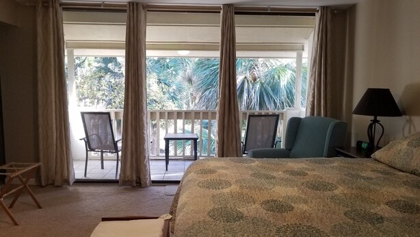 A hotel room with a bed, a desk, a chair, and a balcony with a view of palm trees.