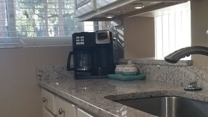 A kitchen with white cabinets, a granite countertop, a coffee maker, a sink, and a window with blinds.