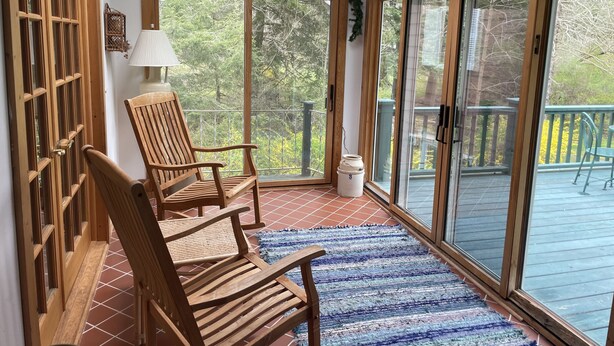 Une chaise en bois et une lampe de table dans une pièce baignée de lumière, avec vue sur une terrasse et des arbres.