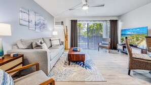 Una sala de estar moderna con sofá, mesa de centro y televisor. Hay un ventilador de techo, una ventana con cortinas y vistas a zonas verdes.