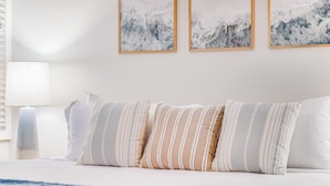 Una cama bien hecha con almohadillas a rayas y una manta azul, una lámpara de noche y tres cuadros abstractos en la pared.