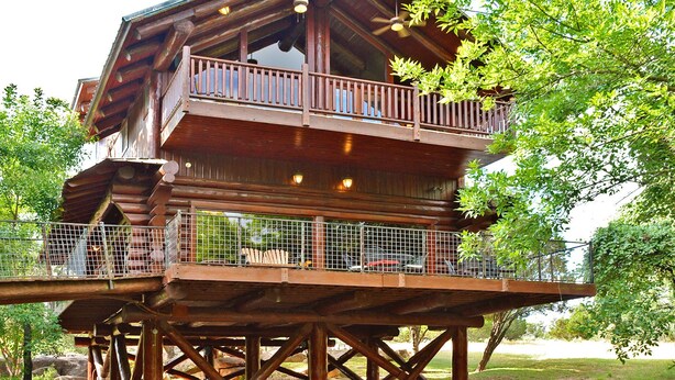A wooden multi-level structure with a balcony, built over a body of water, surrounded by greenery. - Lakefront•3BR/3.5BA+Loft•Hot Tub&Boat House (Burnet)