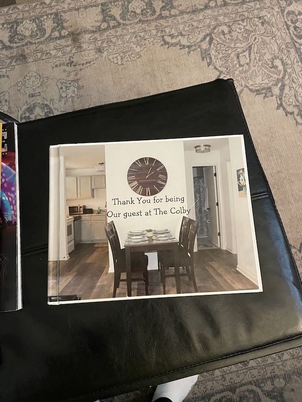 A framed photograph of a dining area with a clock, a message thanking a guest, and a kitchen in the background.