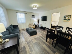 A living room with a sofa, ottoman, dining table with chairs, a flat-screen TV, and a window with blinds.