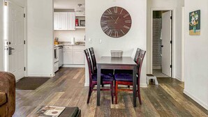 Een eetgedeelte met een houten tafel en stoelen, een grote wandklok en een keuken op de achtergrond.