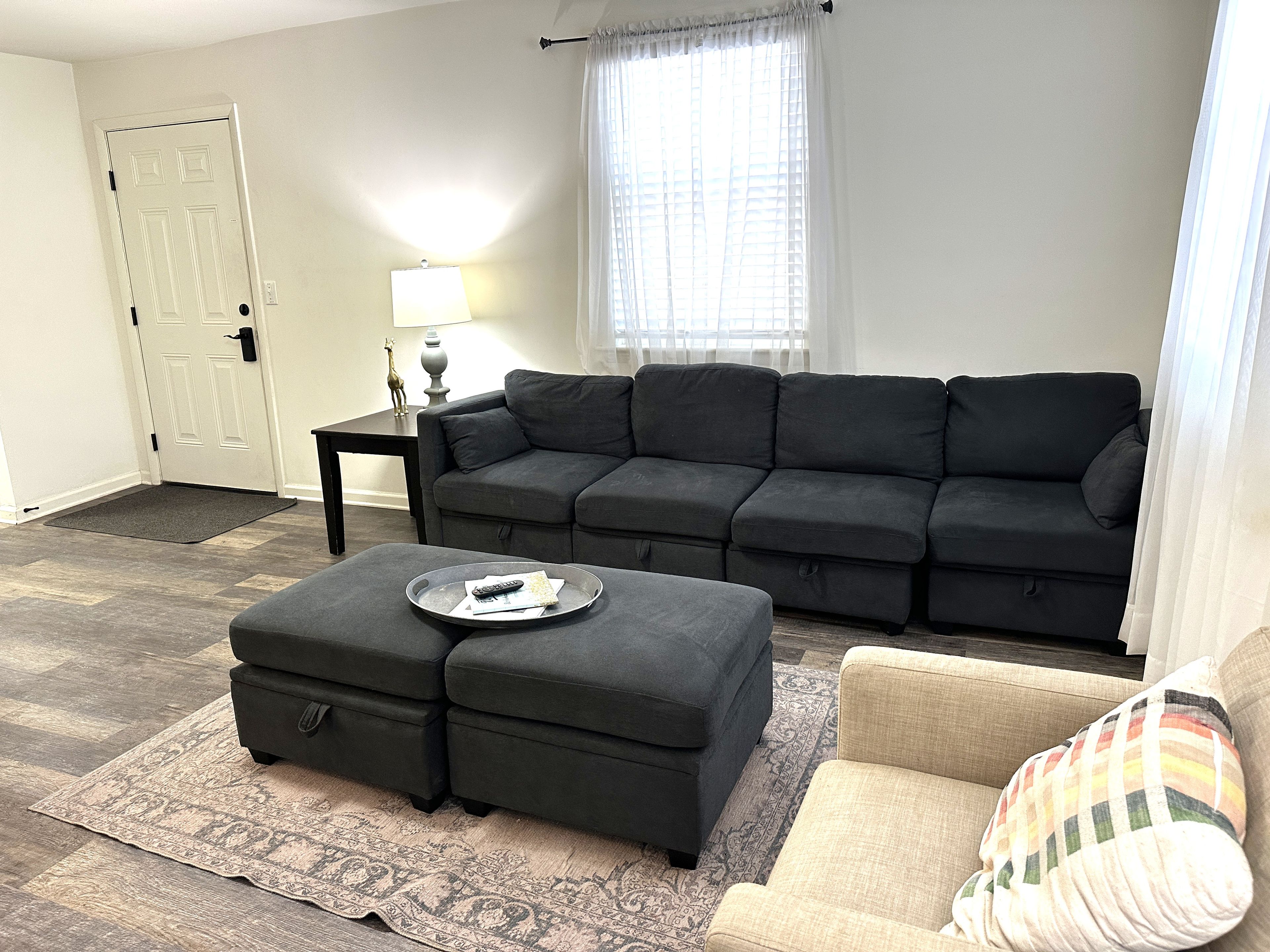 A living room with a sectional sofa, a small table with a lamp, a coffee table with remote controls, and a patterned rug.