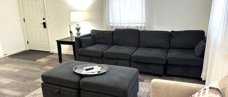 A living room with a sectional sofa, a small table with a lamp, a coffee table with remote controls, and a patterned rug.