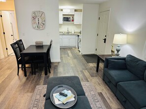 A living room with a dark green sofa, a dining table with chairs, a kitchen area with white cabinets, and a wall clock.
