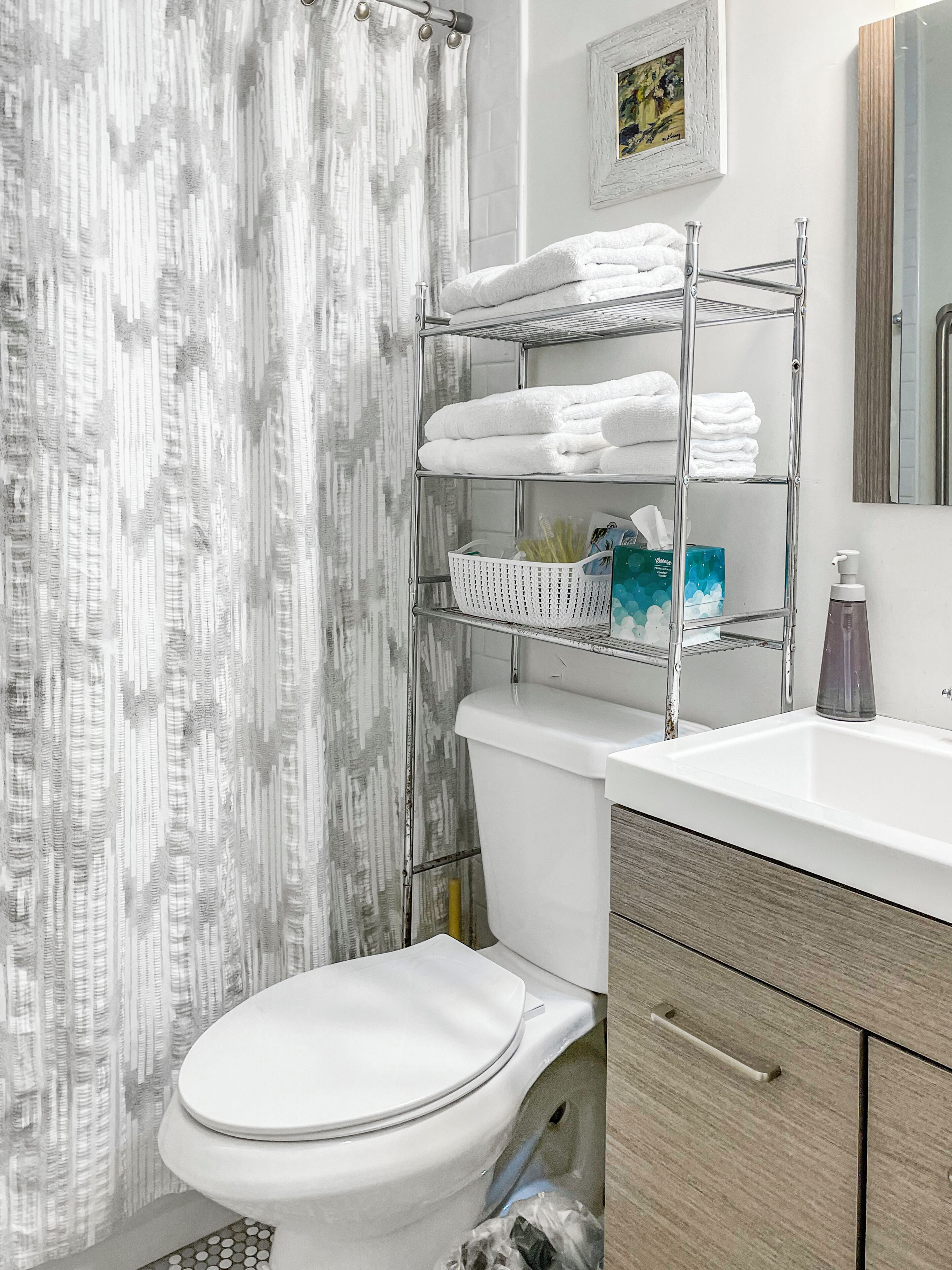 Une salle de bain avec des toilettes blanches, un rideau de douche, un porte-serviettes en métal avec des serviettes pliées, un panier et un miroir.