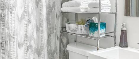 Une salle de bain avec des toilettes blanches, un rideau de douche, un porte-serviettes en métal avec des serviettes pliées, un panier et un miroir.