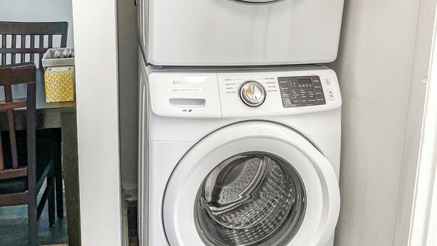 Una lavadora y secadora apiladas en una esquina, con una silla y una cesta de ropa a la vista.
