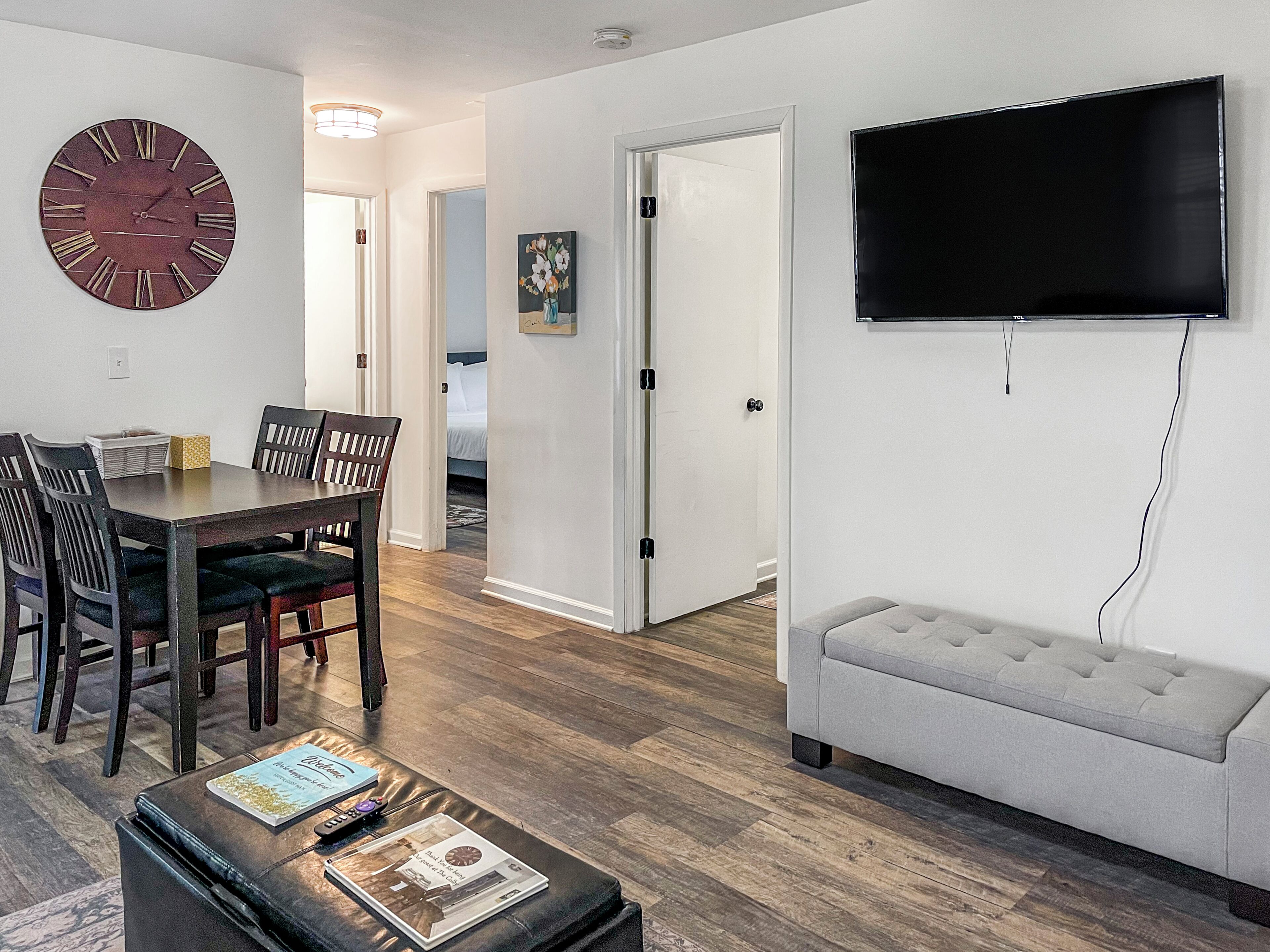A living room with a dining table, chairs, a sofa, a flat-screen TV, and a clock on the wall.