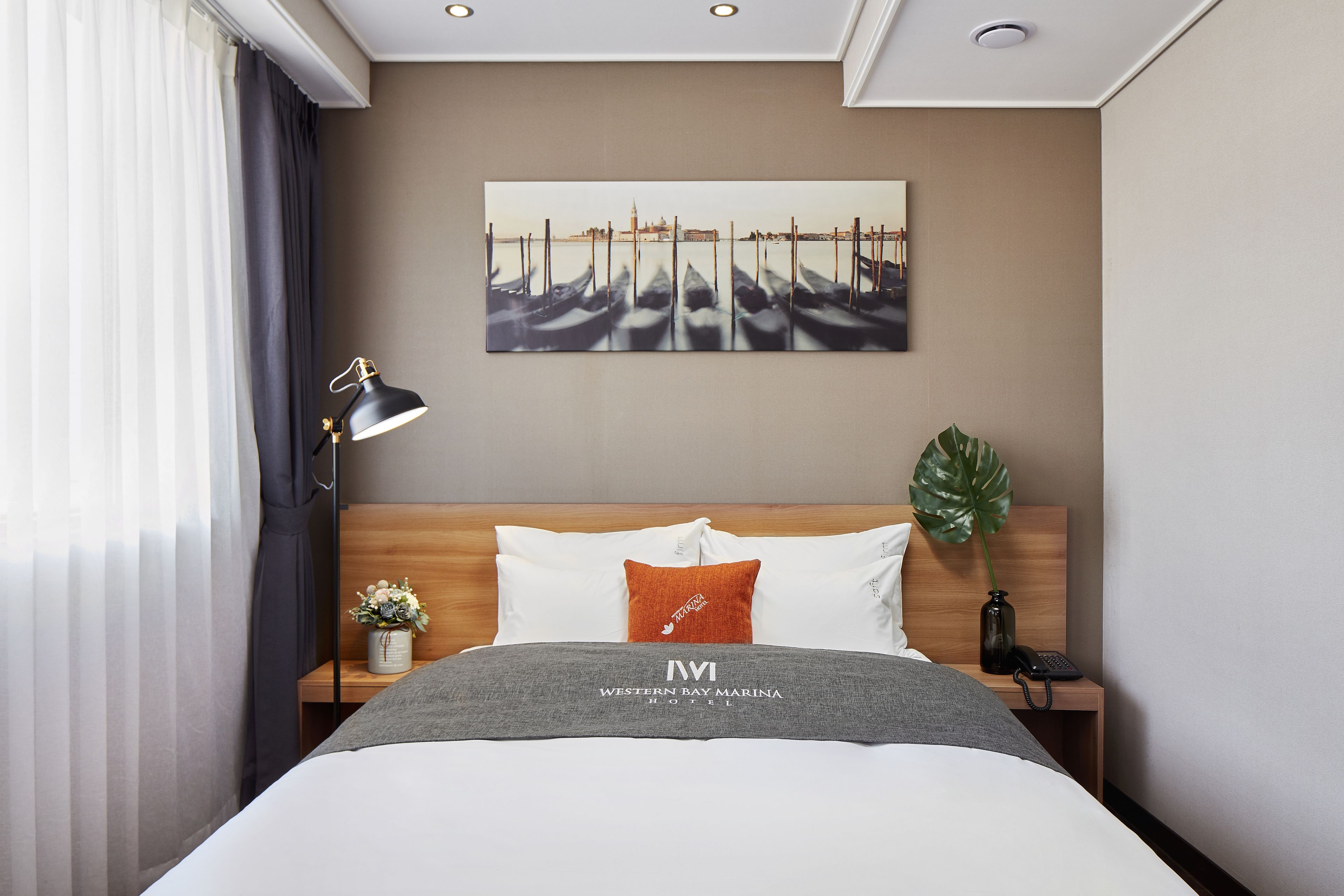 A hotel room with a bed, a nightstand, a lamp, a painting, and a plant.