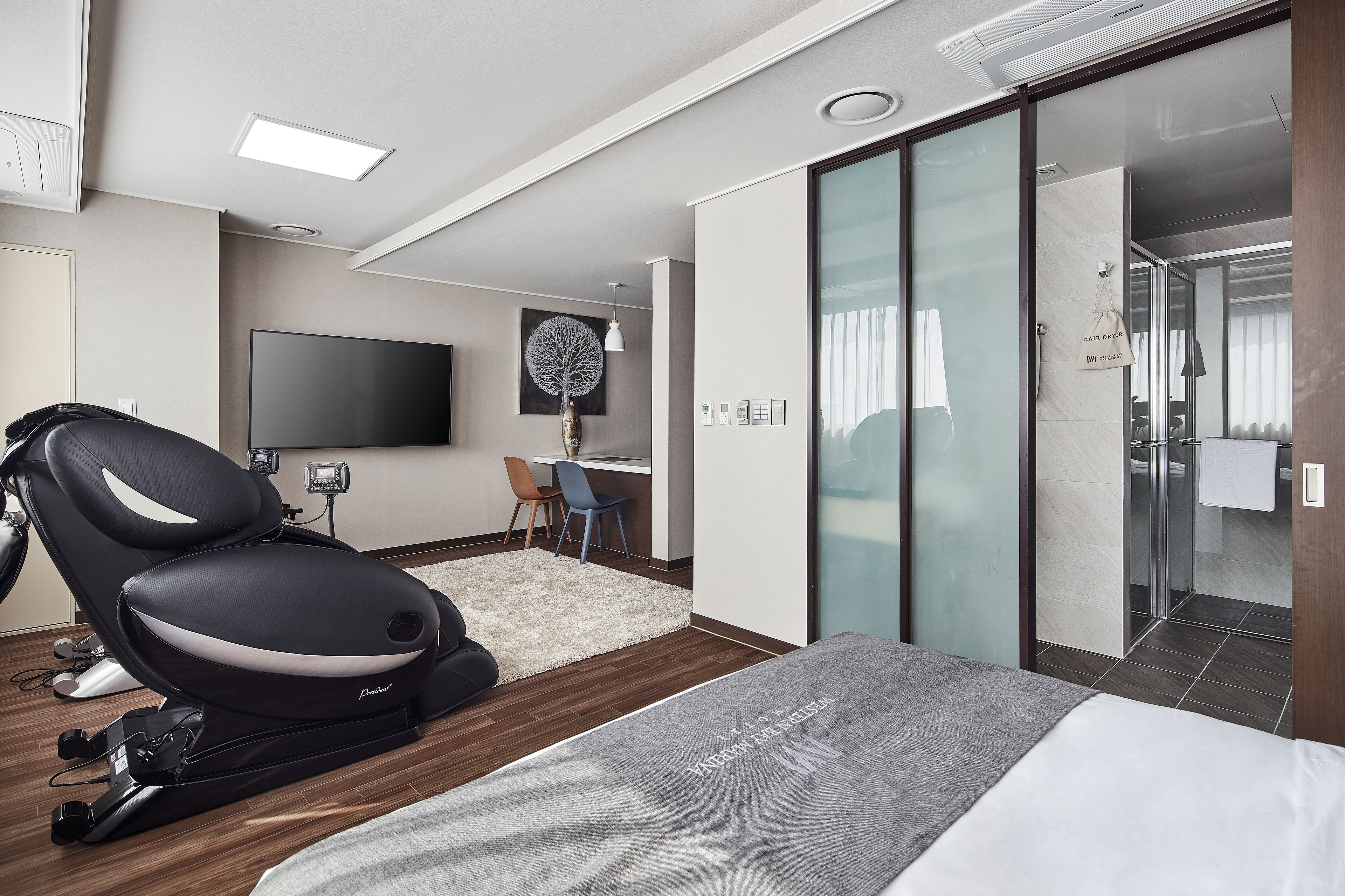 Ett modernt hotellrum med en massagefåtölj, en platt-TV, ett skrivbord med stol och en skjutdörr i glas som leder till badrummet.