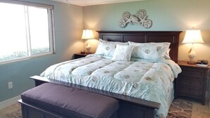 A bedroom with a bed, two bedside tables, a nightstand, a window, and a decorative wall piece. - Summerhouse 101:  Luxury Condo on Beach (St. Augustine)