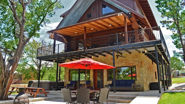 A two-story wooden cabin with a balcony, stone walls, and outdoor seating with a red umbrella.