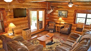 A log cabin interior with leather sofas, a wooden coffee table, a television mounted on the wall, and a mounted deer head.