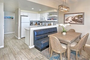 A modern kitchen with white cabinets, a built-in oven, and a central island with a blue bench. There is a dining area with a wooden table and wicker chairs.