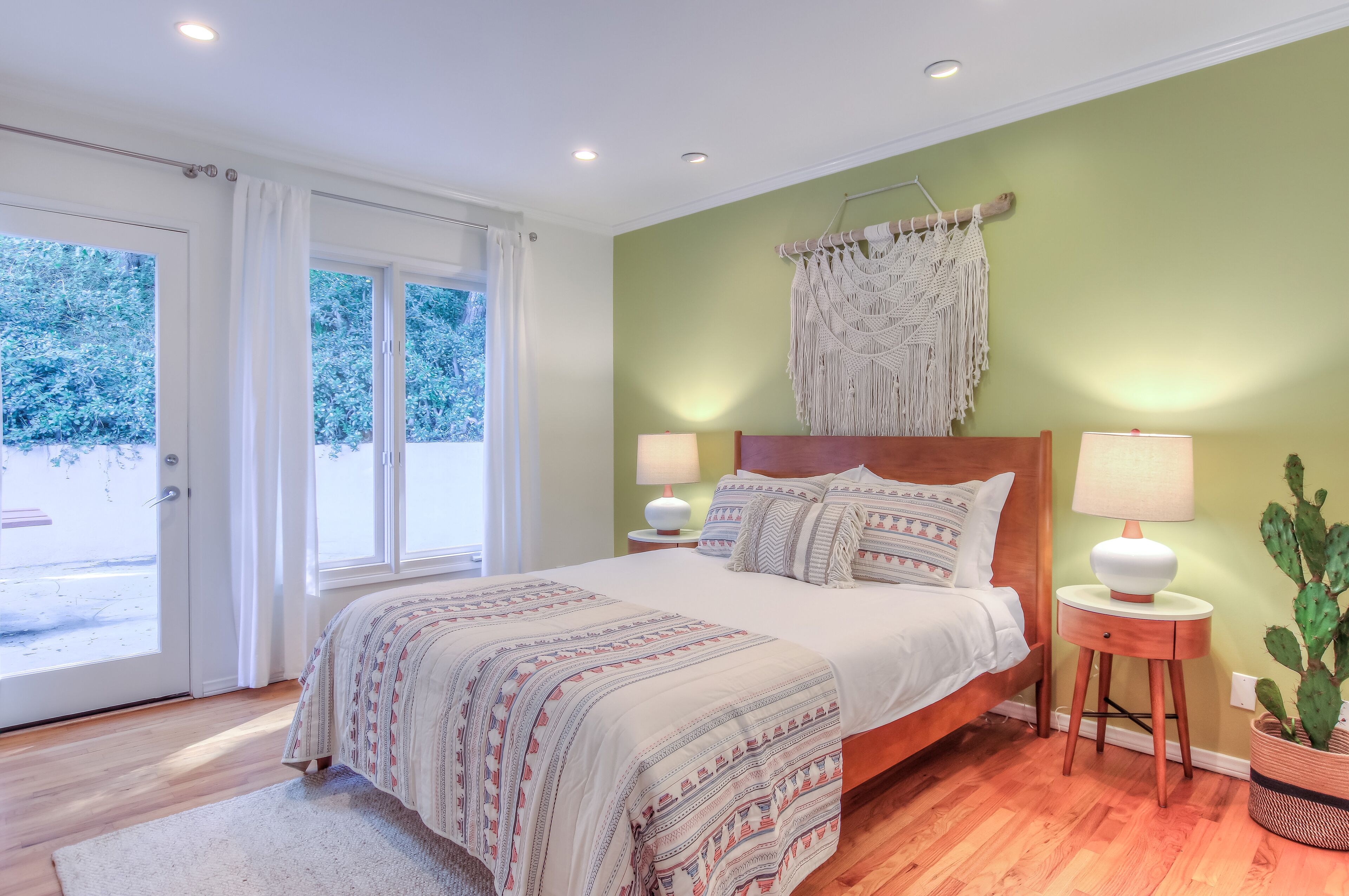 Une chambre avec un lit, deux lampes de chevet, une décoration murale en macramé, un cactus et une vue sur l’extérieur par une porte vitrée.