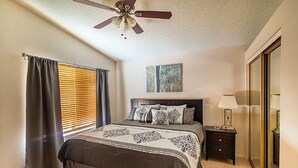 Une chambre à coucher comprenant un lit, une table de chevet, une lampe, un ventilateur de plafond et une fenêtre avec des stores.