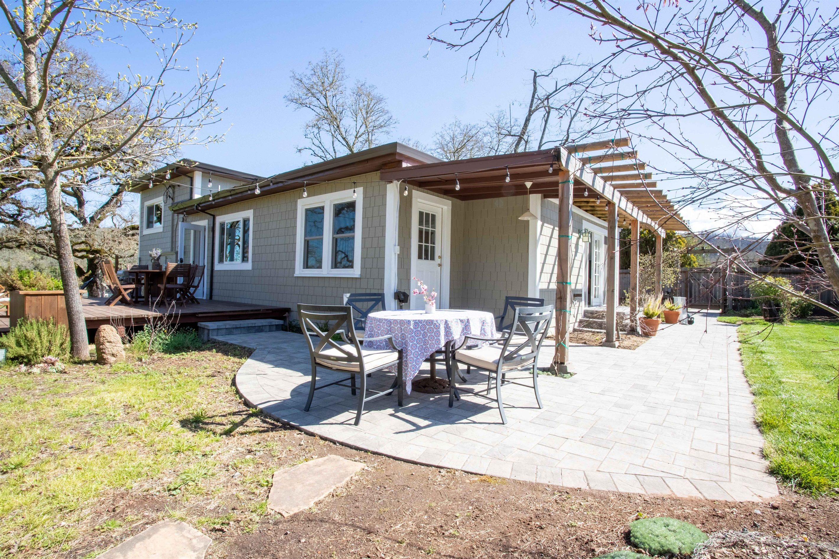 A house with a patio area featuring a table and chairs, a pergola, and a garden with potted plants.