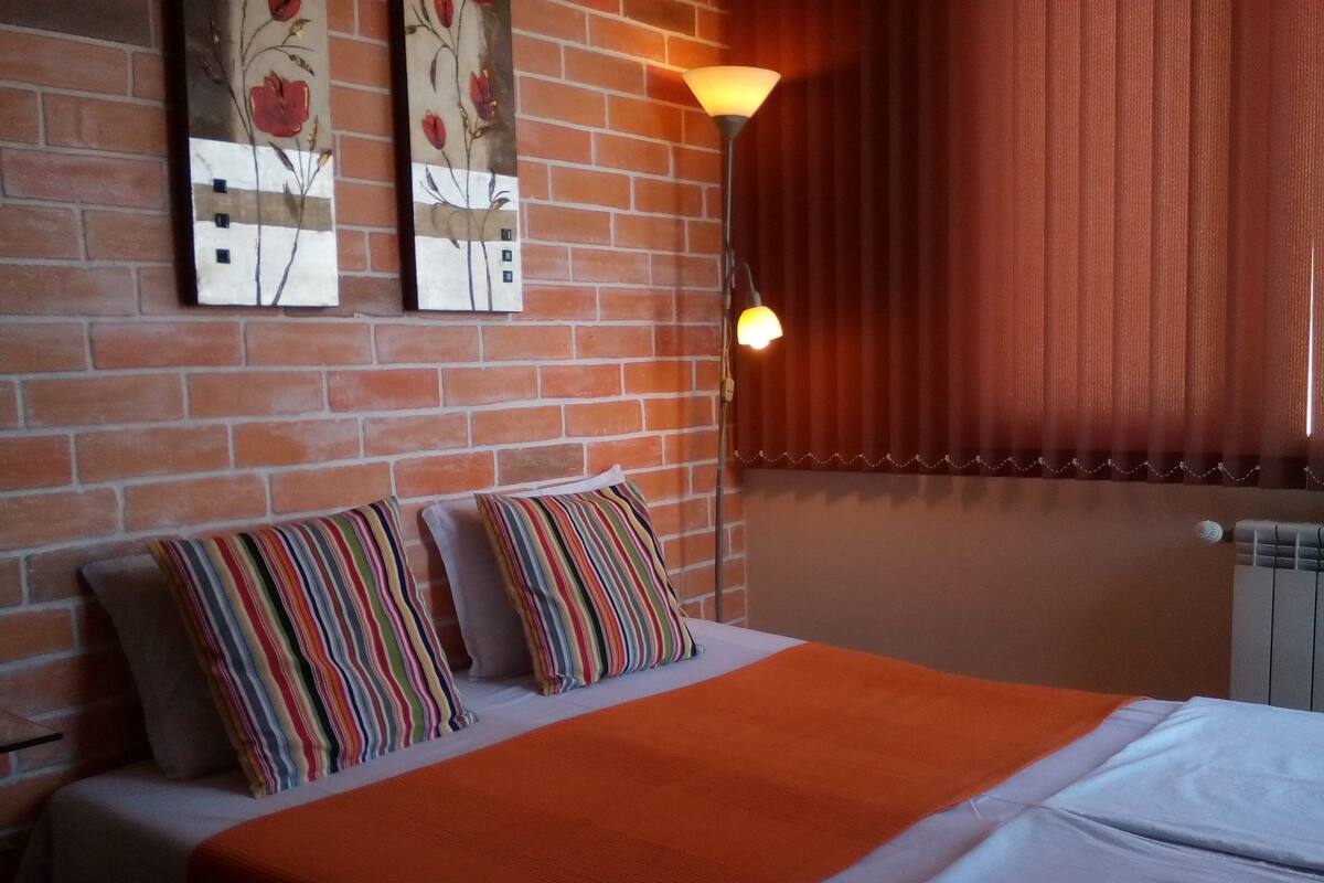 A hotel room with a brick wall, a bed with striped pillows, an orange blanket, and a wall-mounted lamp.