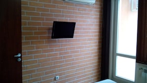 A room with a brick wall, a mounted air conditioner, a flat-screen TV, a window with curtains, and a door with a handle. - Greek Rooms in the City Center (Sofia)